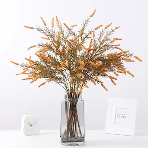 Op de afbeelding: Een boeket gedroogde gele bloemen in een heldere glazen vaas met een grijze tint. De vaas staat op een wit oppervlak met een witte klok en een wit fotolijstje met de tekst "Weekend in Parijs".