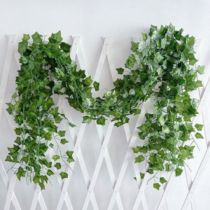 May include: Two strands of artificial green ivy vines hanging on a white lattice fence. The vines are made of plastic and have a realistic look.