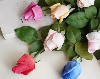 Pode incluir: Uma vista aérea de várias rosas artificiais em várias cores, incluindo rosa, vermelho, amarelo e azul. As rosas estão em diferentes estágios de floração, com folhas e caules verdes. As flores estão dispostas sobre uma superfície branca.