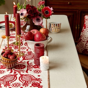 Peut inclure: Une nappe blanche avec un chemin de table brodé rouge, orné d'un motif floral. La table est dressée avec des bougies, un bol de pommes et un bol décoratif.