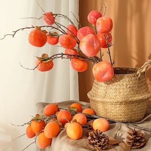 Può includere: Un bouquet di cachi artificiali arancioni su rami in un cesto intrecciato. I cachi sono disposti in un vaso e su un tavolo con pigne.