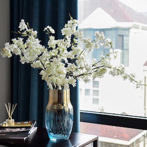 May include: A blue and gold glass vase with a white cherry blossom branch arrangement. The vase is on a black table in front of a window.
