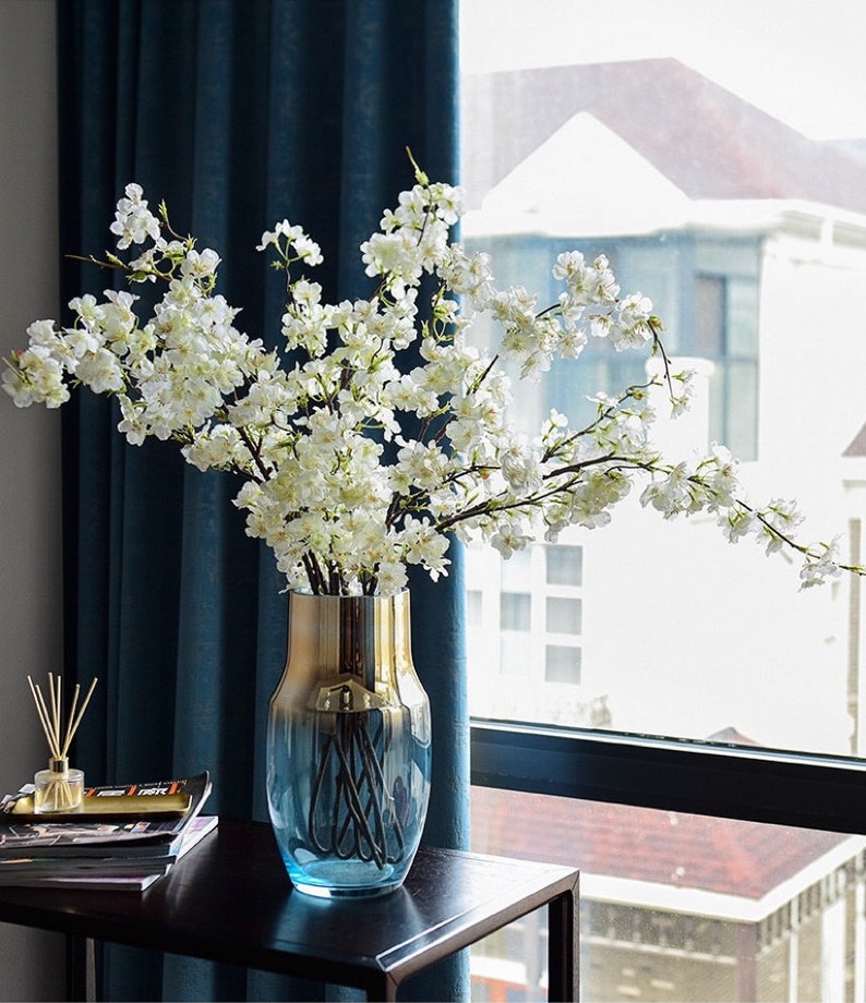 May include: A blue and gold glass vase with a white cherry blossom branch arrangement. The vase is on a black table in front of a window.