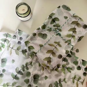 May include: A rectangular gift wrapped in white tissue paper featuring a green eucalyptus leaf pattern, tied with thin green and white twine. Beside the gift lies a matching sheet of eucalyptus tissue paper and a small glass candle jar.