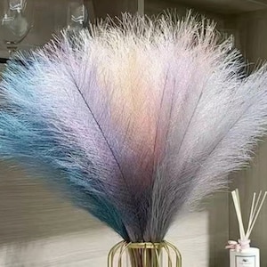 Puede incluir: Un ramo de hierba de la pampa seca en tonos azules, rosas y blancos. La hierba está dispuesta en un jarrón con un soporte de metal dorado.