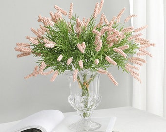 May include: A bouquet of pink artificial lavender flowers in a clear glass vase with a decorative design. The vase is sitting on a white surface with a book.