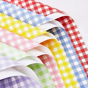 May include: A collection of colorful gingham patterned papers in red, blue, yellow, pink, green, and purple. The papers are arranged in a fan shape, with water droplets visible on the surface. The classic check pattern is a common design element.