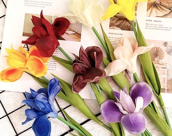 Può includere: Una collezione di sette fiori di iris artificiali in vari colori, tra cui giallo, bianco, rosso scuro, arancione, viola scuro, rosa chiaro e blu. Ogni fiore presenta lunghi steli e foglie verdi, disposti insieme in un vivace bouquet multicolore.
