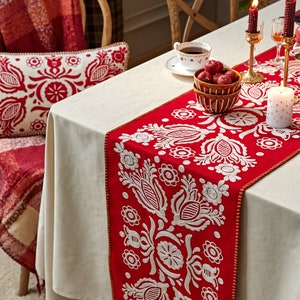 以下が含まれることがあります： 白のテーブルクロスに赤と白の刺繍が施されたテーブルランナーが置かれています。ランナーは花柄のデザインで、茶色の縁取りがされています。テーブルランナーの上に赤いリンゴの入ったボウルが置かれています。