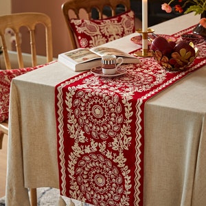 May include: A red and white table runner with a paisley design and tassels. The runner is on a beige tablecloth with a book and a cup of coffee on a saucer.
