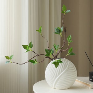 May include: A white ceramic vase with a leaf design holds branches with green leaves. The vase is round and sits on a white table. The branches are brown and have multiple green leaves. The background is a neutral color.