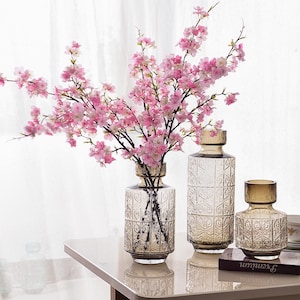May include: Three clear glass vases with an embossed pattern, filled with pink cherry blossom branches. The vases are on a white table with a book.