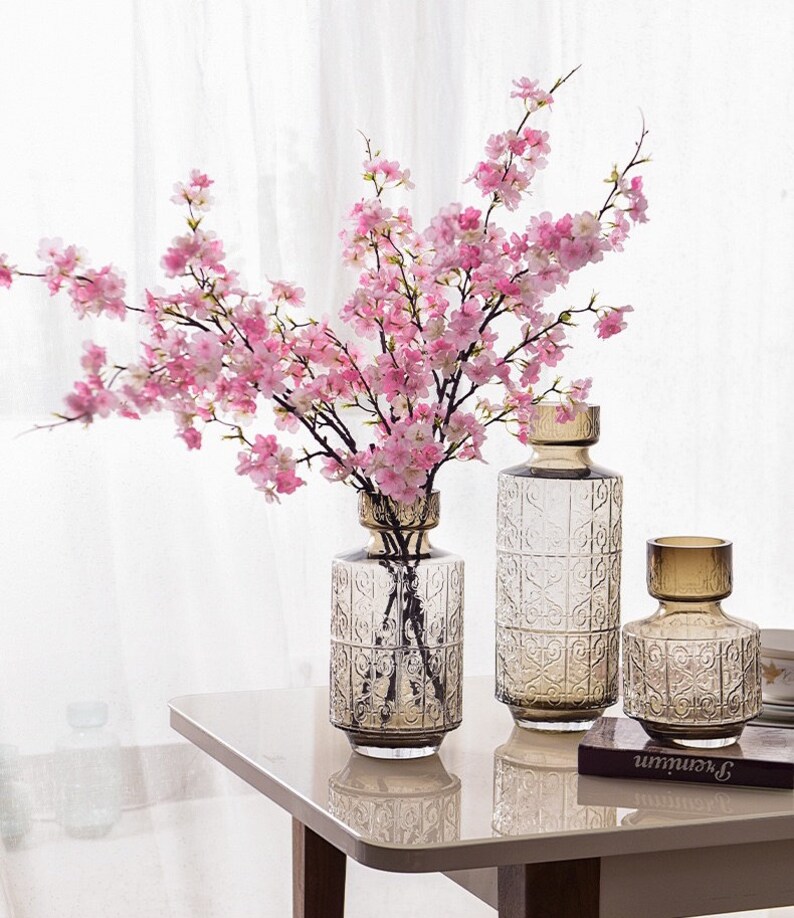 May include: Three clear glass vases with an embossed pattern, filled with pink cherry blossom branches. The vases are on a white table with a book.