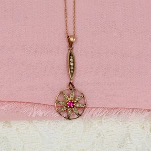 Puede incluir: Un collar con colgante dorado con un diseño circular. Presenta una gema roja central y pequeñas gemas blancas. El colgante cuelga de una delicada cadena, con una sección vertical adornada con pequeñas perlas. El collar descansa sobre una tela rosa.