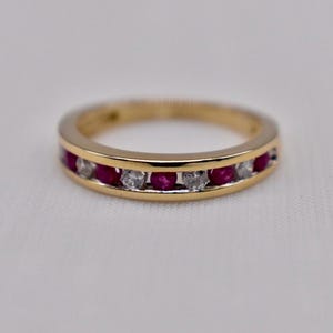 May include: A gold band ring featuring a channel setting with alternating round-cut red rubies and clear white diamonds. The polished yellow gold metal creates a smooth, continuous circular frame around the stones, showcasing a classic design.