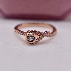 May include: Rose gold ring with a central diamond and smaller accent stones. The ring features a unique knot design, with a round diamond at the center. The band is adorned with small, sparkling stones. A pink jewelry box is in the background.