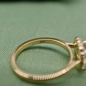 K&ouml;nnte beinhalten: Ein goldener Ring mit einem Halo-Besatz aus kleinen Diamanten. Der Ring ist auf der Innenseite der Ringschiene mit dem Text "14K" graviert.