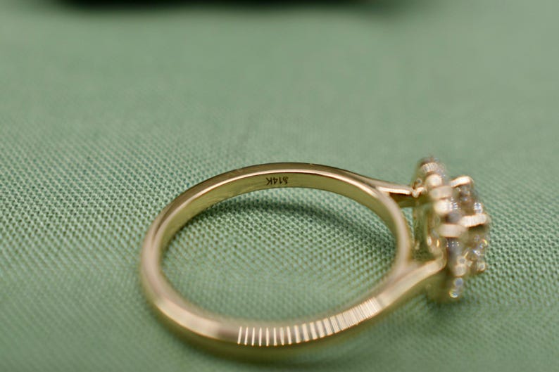 K&ouml;nnte beinhalten: Ein goldener Ring mit einem Halo-Besatz aus kleinen Diamanten. Der Ring ist auf der Innenseite der Ringschiene mit dem Text "14K" graviert.