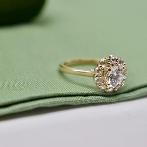 K&ouml;nnte beinhalten: Ein goldener Verlobungsring mit einem gro&szlig;en runden Diamanten, der von kleineren Diamanten umgeben ist. Der Ring liegt auf einem gr&uuml;nen Stoffhintergrund.