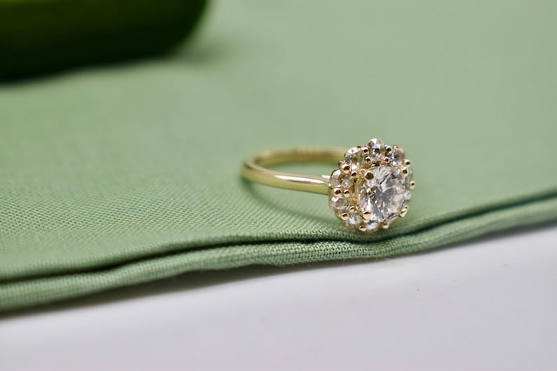 K&ouml;nnte beinhalten: Ein goldener Verlobungsring mit einem gro&szlig;en runden Diamanten, der von kleineren Diamanten umgeben ist. Der Ring liegt auf einem gr&uuml;nen Stoffhintergrund.
