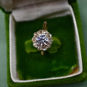 K&ouml;nnte beinhalten: Ein goldener Ring mit einem gro&szlig;en, runden, klaren Diamanten, der von kleineren Diamanten umgeben ist. Der Ring befindet sich in einer gr&uuml;nen Samt-Ringbox.