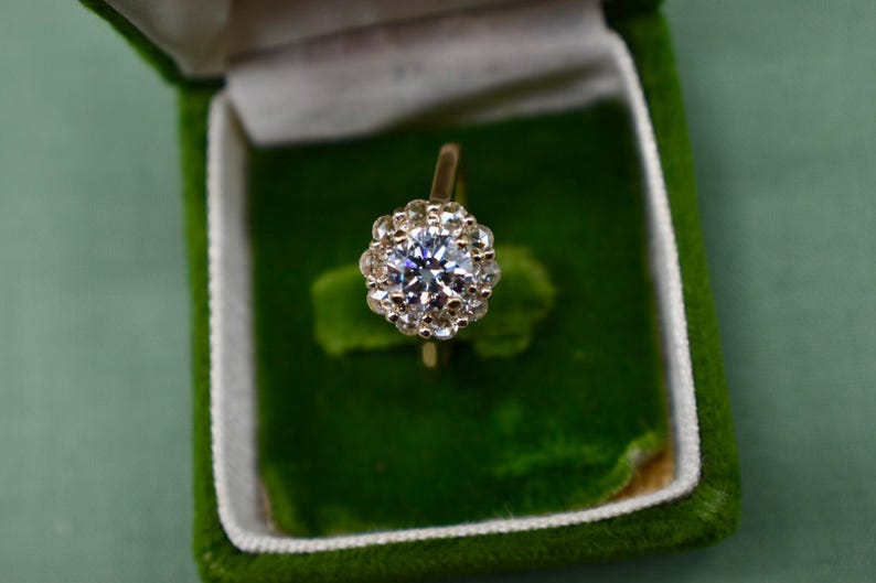 K&ouml;nnte beinhalten: Ein goldener Ring mit einem gro&szlig;en, runden, klaren Diamanten, der von kleineren Diamanten umgeben ist. Der Ring befindet sich in einer gr&uuml;nen Samt-Ringbox.
