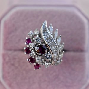 Puede incluir: Un anillo de tono plateado con un grupo de rubíes rojos de corte redondo y diamantes blancos transparentes. El diseño incluye una fila curva de diamantes de corte baguette junto a diamantes de corte marquesa y redondo en una banda metálica.