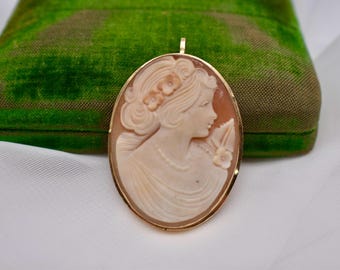 May include: Oval cameo pendant with a profile of a woman in a gold setting. The cameo is carved from a light peach-colored material, with a gold-colored frame. The background is a green velvet box and white fabric.