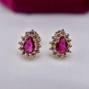 May include: A pair of gold-toned stud earrings. Each earring features a teardrop-shaped, vibrant pink gemstone surrounded by a halo of smaller, clear gemstones. The earrings are set against a white background.