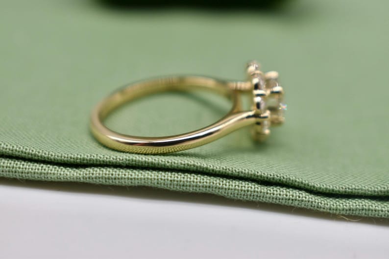 K&ouml;nnte beinhalten: Ein goldener Verlobungsring mit einem runden Mittelst&uuml;ck, das von kleineren Steinen umgeben ist. Der Ring liegt auf einem gr&uuml;nen Stoffhintergrund.