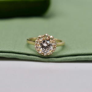 K&ouml;nnte beinhalten: Ein goldener Verlobungsring mit einem runden Diamanten in der Mitte, umgeben von einem Heiligenschein aus kleineren Diamanten.