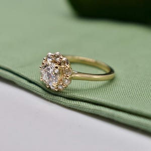 K&ouml;nnte beinhalten: Ein goldener Ring mit einem gro&szlig;en, runden, klaren Edelstein, der von kleineren, klaren Edelsteinen umgeben ist. Der Ring liegt auf einem gr&uuml;nen Stoffhintergrund.