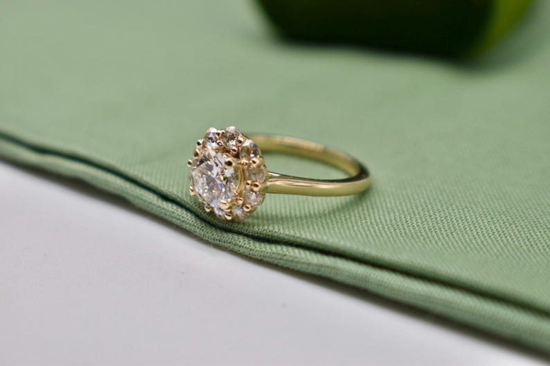 K&ouml;nnte beinhalten: Ein goldener Ring mit einem gro&szlig;en, runden, klaren Edelstein, der von kleineren, klaren Edelsteinen umgeben ist. Der Ring liegt auf einem gr&uuml;nen Stoffhintergrund.