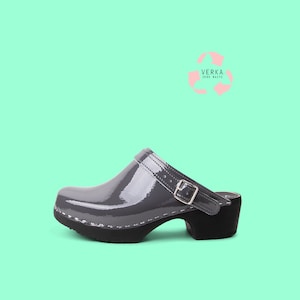 May include: Gray patent leather clog with a black buckle and a black heel. The clog is on a light green background. VERKA. ZERO WASTE is written in black text on a pink recycling symbol.