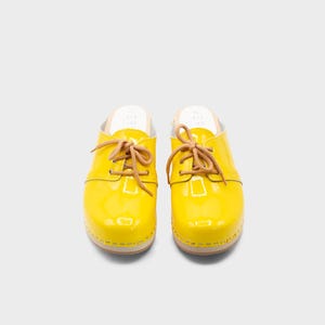 May include: Pair of bright yellow clogs with a glossy finish. The clogs feature tan-colored laces and a wooden sole. The shoes are designed in a classic style, suitable for casual wear. The clogs are a vibrant addition to any wardrobe.