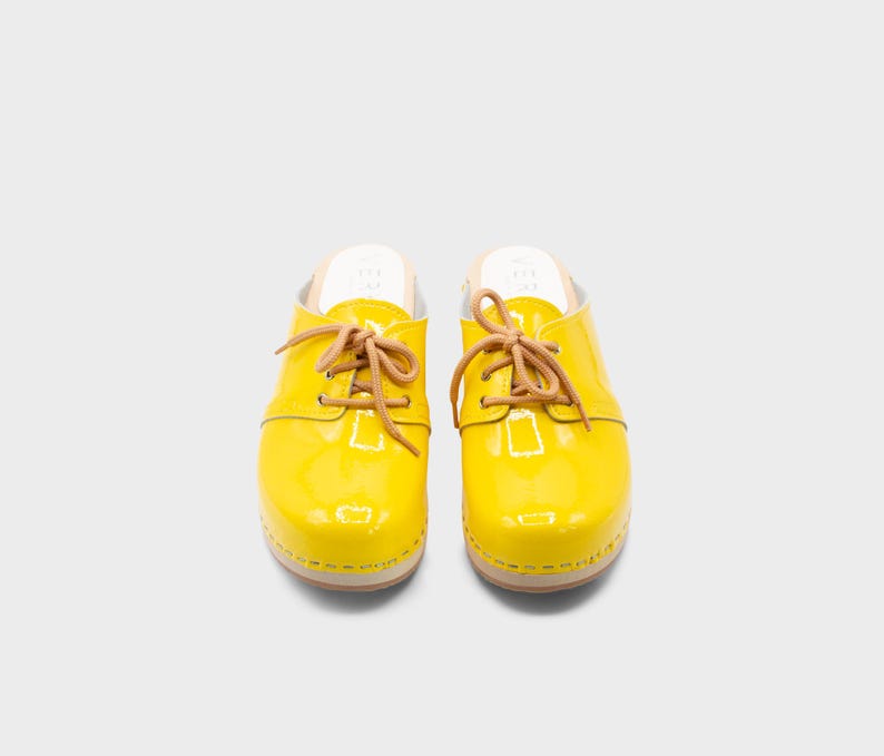 May include: Pair of bright yellow clogs with a glossy finish. The clogs feature tan-colored laces and a wooden sole. The shoes are designed in a classic style, suitable for casual wear. The clogs are a vibrant addition to any wardrobe.