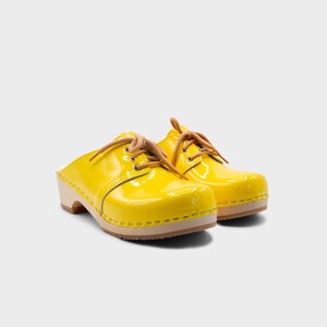 May include: A pair of bright yellow clogs with a glossy finish. The shoes have light brown laces and a wooden sole. The clogs are a classic style, perfect for casual wear.