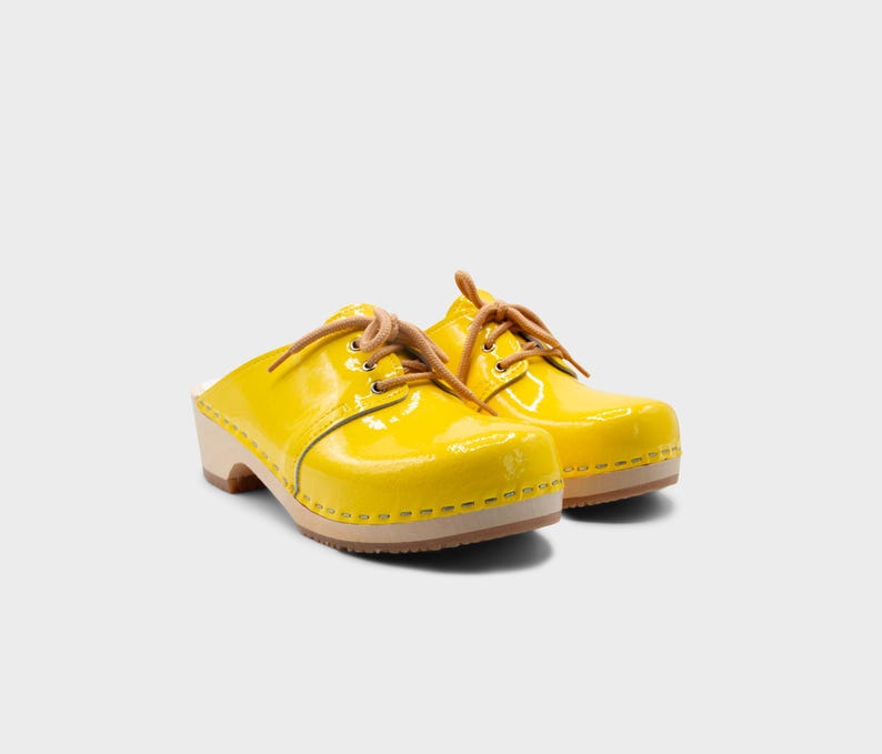 May include: A pair of bright yellow clogs with a glossy finish. The shoes have light brown laces and a wooden sole. The clogs are a classic style, perfect for casual wear.
