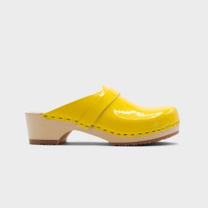Peut inclure: Un sabot jaune vif avec une finition brillante. La chaussure a une semelle et un talon en bois, avec une bride sur le dessus. La semelle mesure environ 2,5 cm d'&eacute;paisseur. Sur fond blanc.