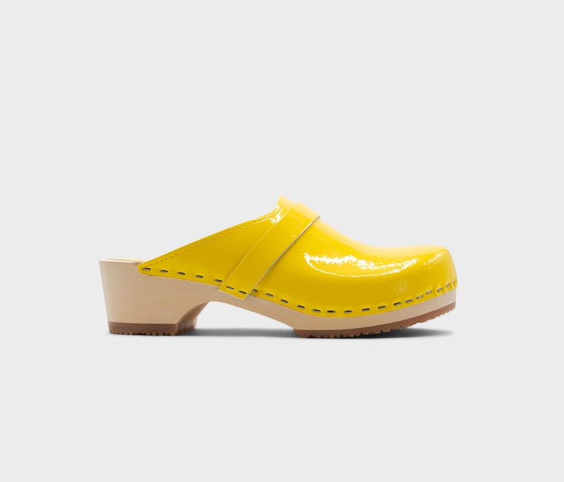 Peut inclure: Un sabot jaune vif avec une finition brillante. La chaussure a une semelle et un talon en bois, avec une bride sur le dessus. La semelle mesure environ 2,5 cm d'&eacute;paisseur. Sur fond blanc.