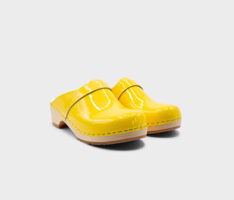 Peut inclure: Une paire de sabots jaune vif avec une finition brillante. Les sabots ont une semelle en bois et une bride sur le dessus. Les chaussures sont d'une couleur vive et ensoleill&eacute;e, parfaites pour un look d&eacute;contract&eacute; et &eacute;l&eacute;gant.