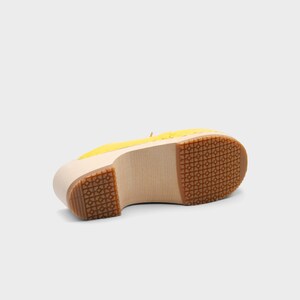 May include: A yellow clog shoe with a wooden sole and a rubber tread. The shoe is shown from the bottom, revealing the light-colored wood and the patterned rubber sole. The upper part of the shoe is yellow.