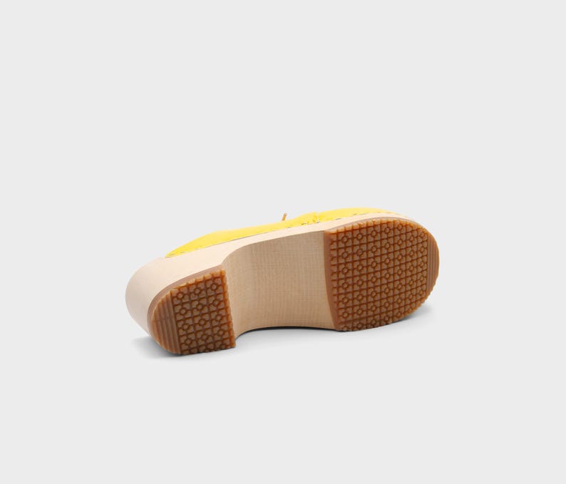 May include: A yellow clog shoe with a wooden sole and a rubber tread. The shoe is shown from the bottom, revealing the light-colored wood and the patterned rubber sole. The upper part of the shoe is yellow.