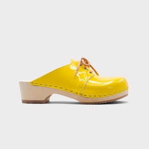 May include: A bright yellow clog with a glossy finish. The shoe features a wooden sole and heel, with a tan leather lace-up detail. The clog is a slip-on style, perfect for casual wear.