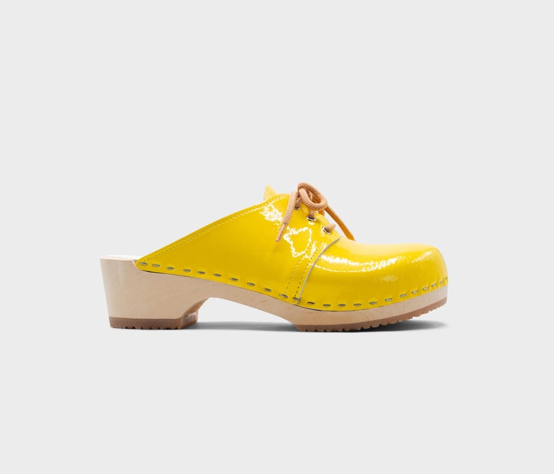 May include: A bright yellow clog with a glossy finish. The shoe features a wooden sole and heel, with a tan leather lace-up detail. The clog is a slip-on style, perfect for casual wear.