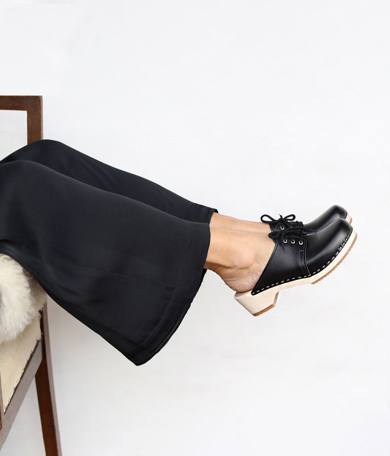 Girl wearing black pants and low heel clog mules in black leather with lace on a light wood base