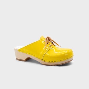 May include: A bright yellow clog with a glossy finish. The shoe features a wooden sole, a rounded toe, and tan-colored laces. The heel is approximately 3 inches high.