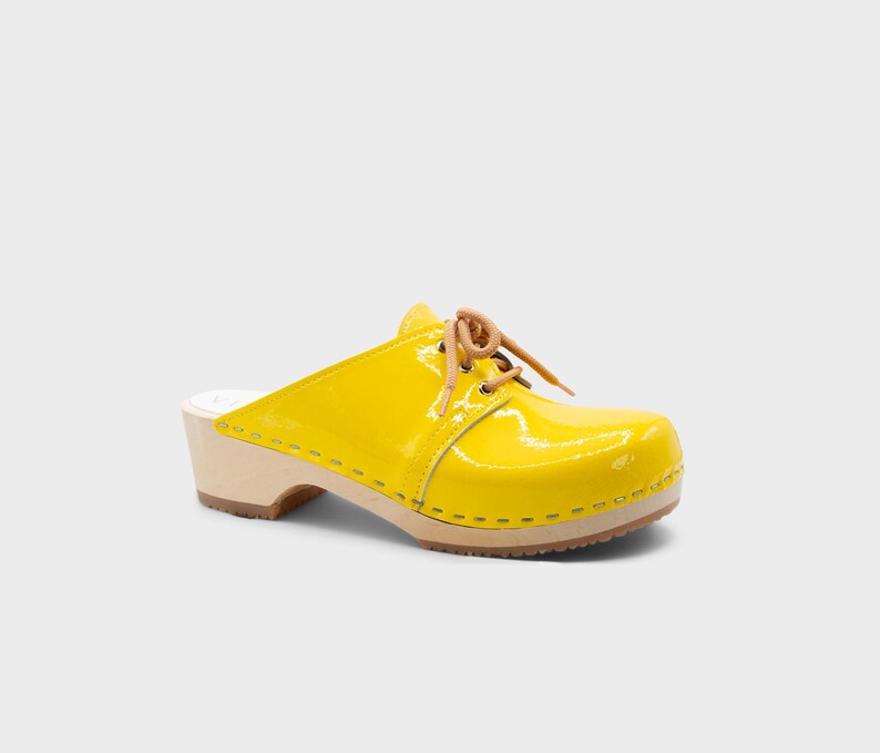May include: A bright yellow clog with a glossy finish. The shoe features a wooden sole, a rounded toe, and tan-colored laces. The heel is approximately 3 inches high.