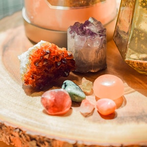 May include: A collection of various crystals and gemstones on a wooden surface. The crystals include a large orange citrine, a purple amethyst, a green aventurine, a pink rose quartz, and a clear quartz.