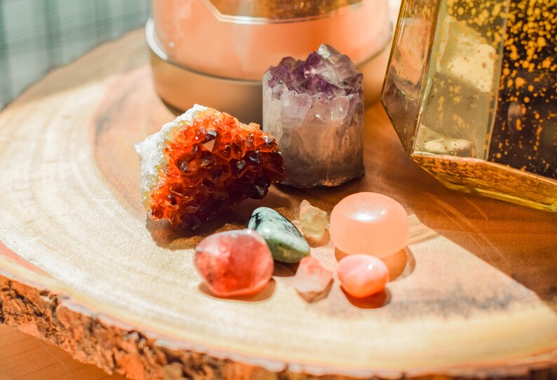 May include: A collection of various crystals and gemstones on a wooden surface. The crystals include a large orange citrine, a purple amethyst, a green aventurine, a pink rose quartz, and a clear quartz.